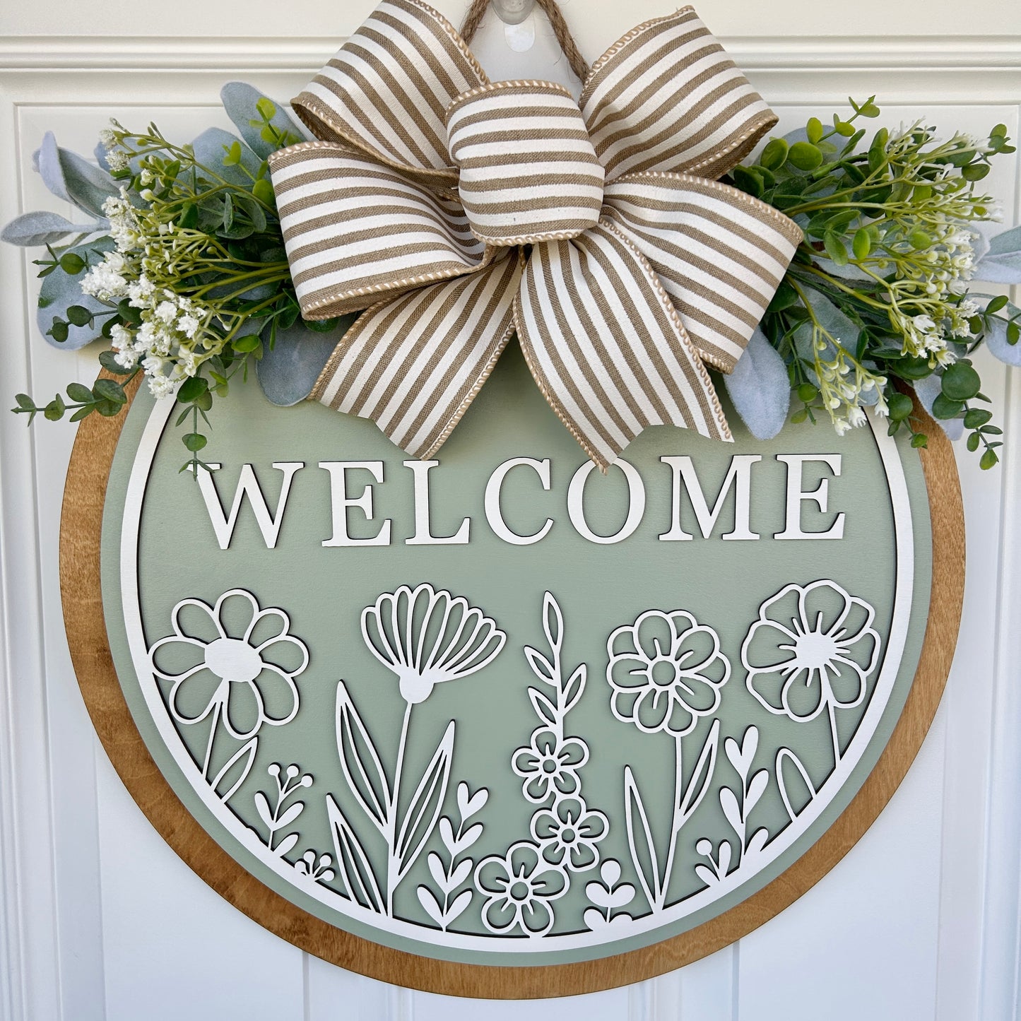 Decorative 'Welcome' sign with white floral designs and a striped bow on a sage background.